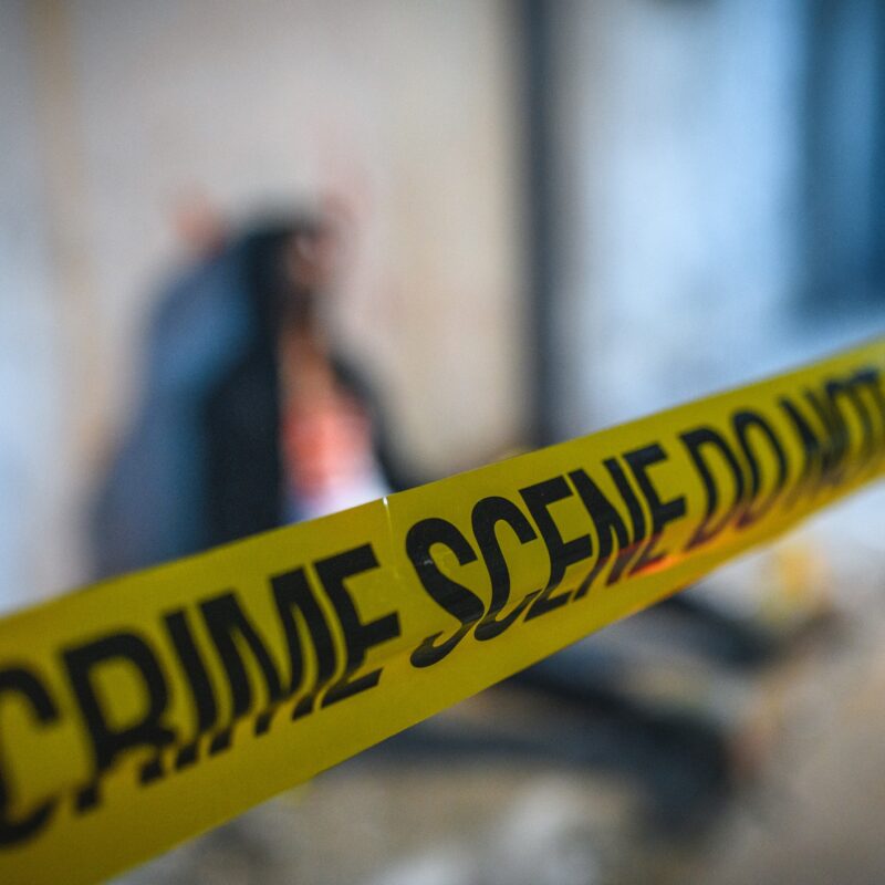 Barricade tape covering a crime scene. There is a dead male body in the background indicating homicide.